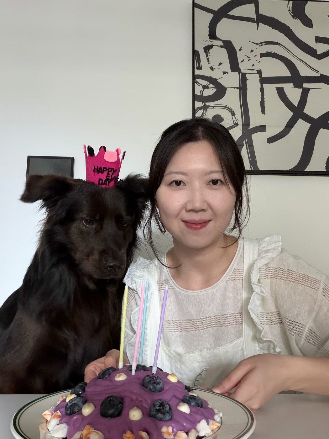 A woman and the same black dog behind a small purple cake with unlit candles; the dog is wearing a pink paper crown.